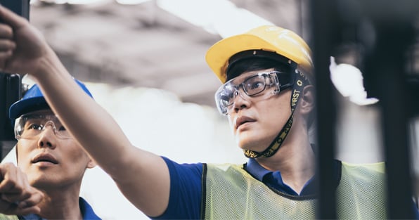 A factory worker in uniform pointing upwards while another factory worker looks in that direction.